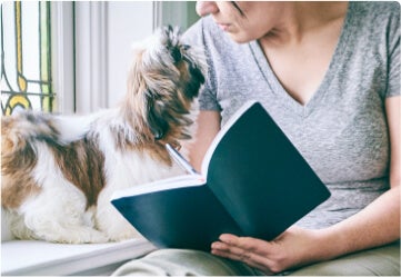 Femme prenant des notes de cours pédagogiques dans un cahier tout en regardant son chien à côté d'elle.